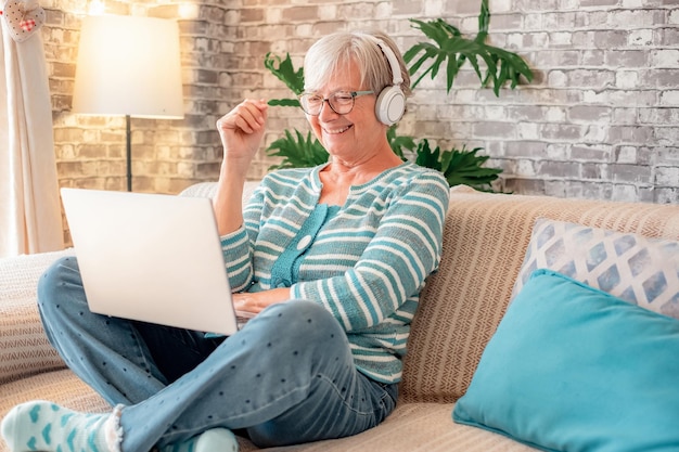 Smiling caucasian senior woman sitting on sofa at home using laptop watching a video