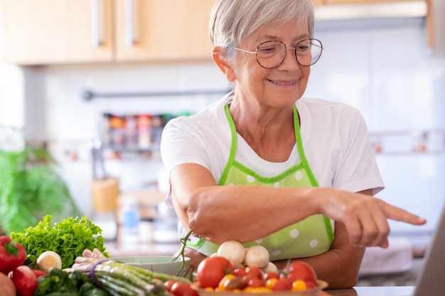 Smiling caucasian senior woman surfing the net sitting in the\
home kitchen while preparing vegetables mature woman using laptop\
technology looking for new recipes