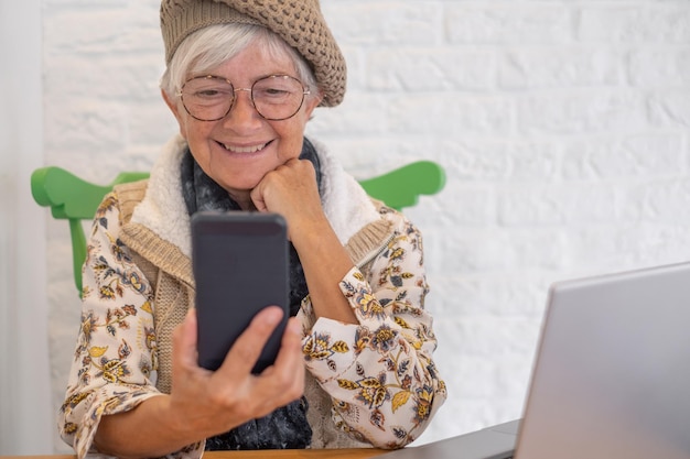 Smiling caucasian senior woman with cap and eyeglasses using mobile phone sitting at cafe shop