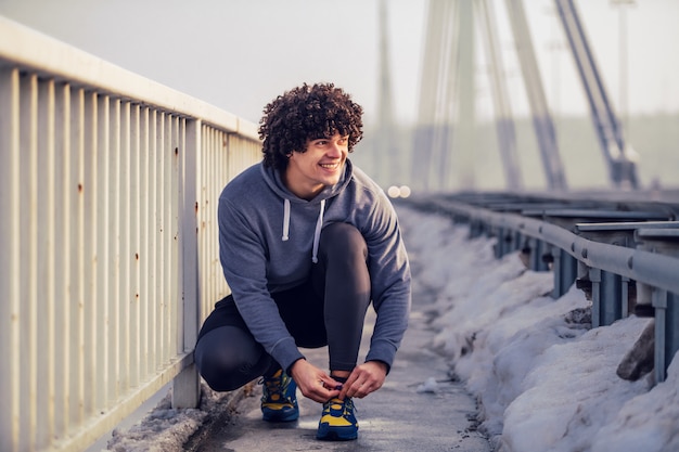 Smiling caucasian sporty man in sportswear and with curly hair crouching on the bridge and tying shoelace. Wintertime. Outdoor fitness concept.