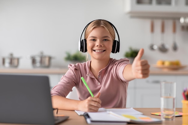 Smiling caucasian teen girl blonde in headphones show thumb up study at home at table with laptop in