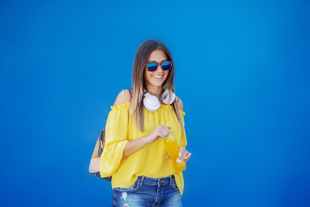 Smiling Caucasian teenage girl in yellow blouse and with sunglasses holding orange juice while standing in front of blue wall.