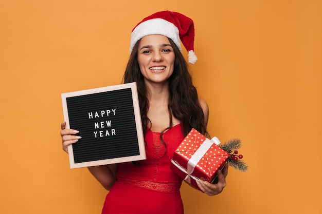 A smiling Caucasian woman in a red Santa hat holding a gift box and a letter board with the inscription Happy New Year on it. On a yellow isolated background.