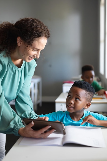 Photo smiling caucasian young female teacher showing digital tablet to african american elementary boy