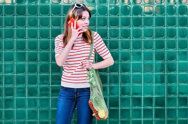 Smiling Caucasian young girl talking on the mobile phone with a net bag over her shoulder with vegetables on a green wall in the street