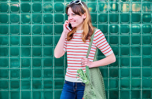 Smiling Caucasian young girl talking on the mobile phone with a net bag over her shoulder with vegetables on a green wall in the street