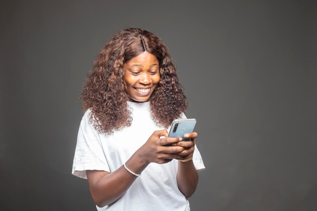 Smiling charming black african woman with afro hairstyle using cellphone standing over a grey studio background chatting with friends on social media