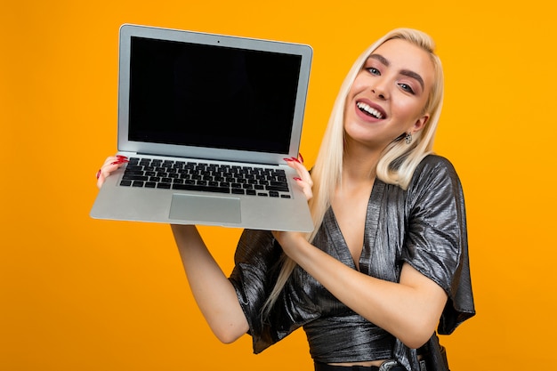 Smiling charming blond girl holding a laptop screen blank on a yellow wall space