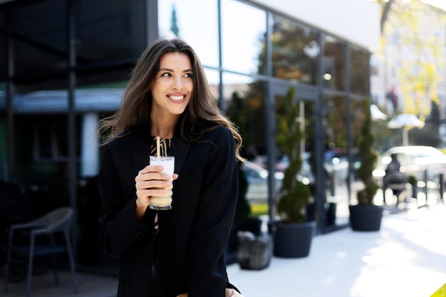 Smiling charming girl with a coffee in her hands is standing on the street looking away The girl stands against the background of a modern glass building