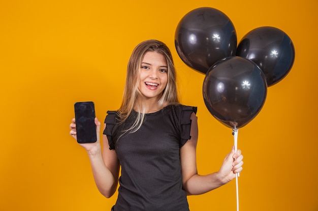 Smiling charming young woman holding a cellphone with empty blank screen on yellow background with air balloons studio portrait. Black Friday settlement