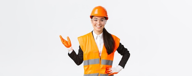 Smiling cheerful asian female construction manager introduce her interprise for investors wearing safety glasses helmet and gloves pointing hand left and looking proud white background