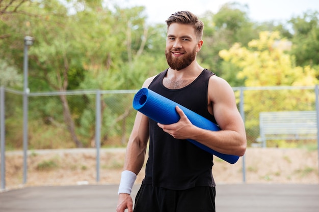 屋外に立っているヨガマットと笑顔の陽気なひげを生やしたフィットネス男