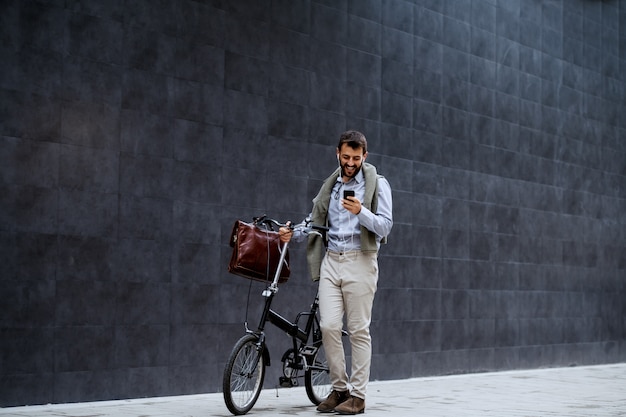 Усмехаясь жизнерадостный кавказский модный бизнесмен используя умный телефон и нажимающ его велосипед. На заднем плане - серая стена.