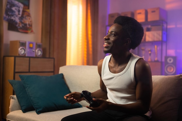 A smiling cheerful darkskinned man sits on a couch in the living room holding a game pad