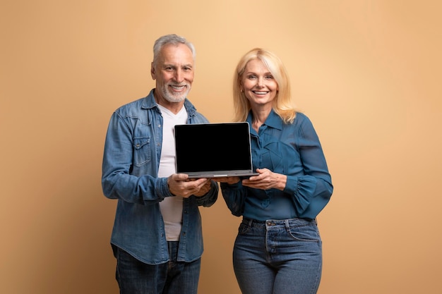 Smiling cheerful funny elderly couple showing laptop with blank screen