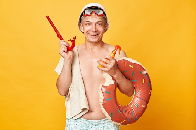 Smiling cheerful positive tourist man at summer resort standing with rubber ring isolated over yellow background looking at camera with happy face holding snorkel