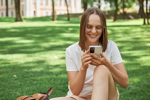 Smiling cheerful positive woman wearing casual white tshirt sitting in park on green grass using mobile phone chatting with friends checking social networks