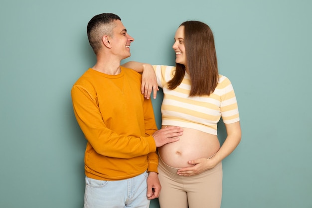 Фото Улыбающаяся веселая беременная пара в повседневной одежде изолирована на синем фоне мужчина и женщина обнимают друг друга и смотрят друг на друга с любовью