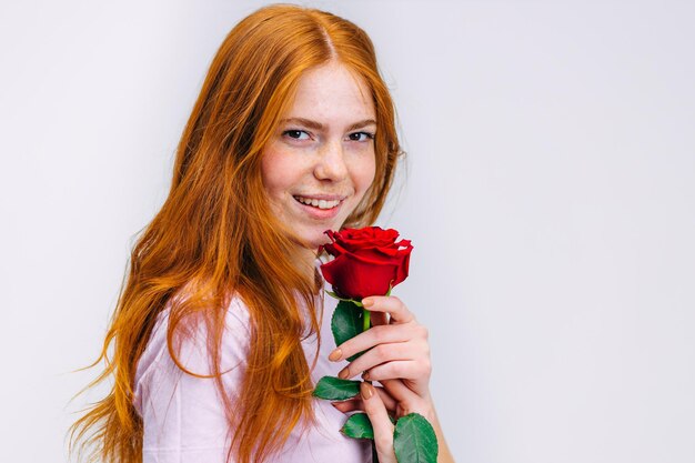 Smiling cheerful redhead girl in white pink tshirt with rose isolated on gray background in studio