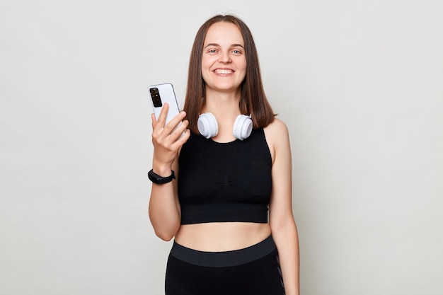 Smiling cheerful slim woman with headphones wearing sportswear posing against gray background using mobile phone sporty app