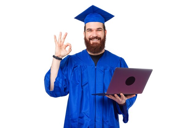 Smiling and cheerful student man in blue robe showing ok and holding laptop