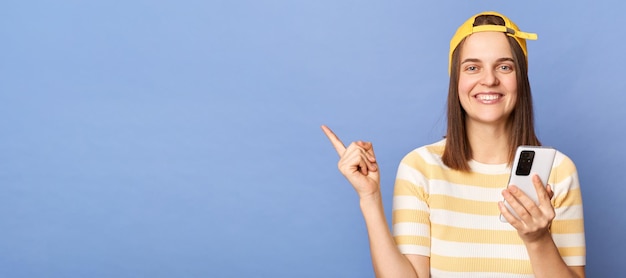 Улыбающаяся веселая девочка-подросток в полосатой футболке и бейсболке, стоящая изолированно на синем фоне, держа смартфон, указывая на рекламную зону