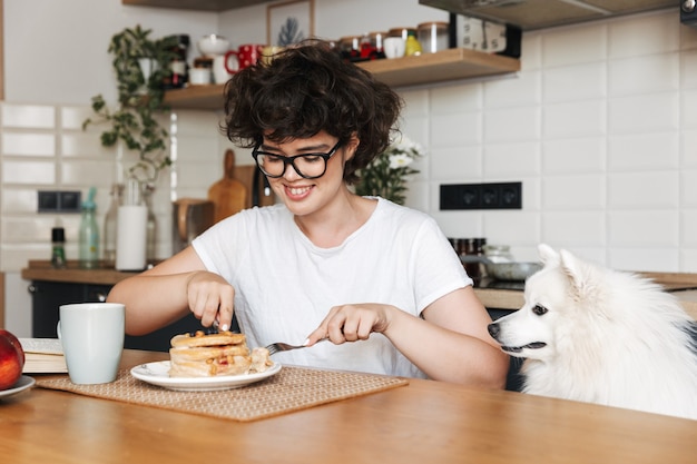 自宅のキッチンに座って、パンケーキを食べながらおいしい朝食を食べて陽気な女性の笑顔