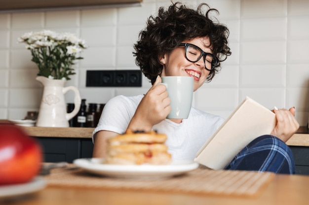 自宅のキッチンに座って、本を読んでおいしい朝食を食べている陽気な女性の笑顔