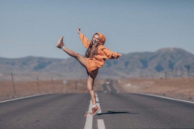 Smiling cheerful woman in orange hoodie poses provocatively with her arm and leg raised on empty highway against the backdrop of a mountain landscape