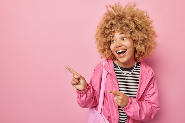 Smiling cheerful woman points index fingers away on left side
demonstrates information banner shows advertisement dressed in
windbreaker isolated over pink background place your promo logo
there