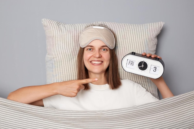 Улыбающаяся жизнерадостная женщина в белой футболке и спящей маске для глаз лежит в постели на подушке под одеялом, изолированная на сером фоне, указывая на будильник, просыпаясь утром вовремя