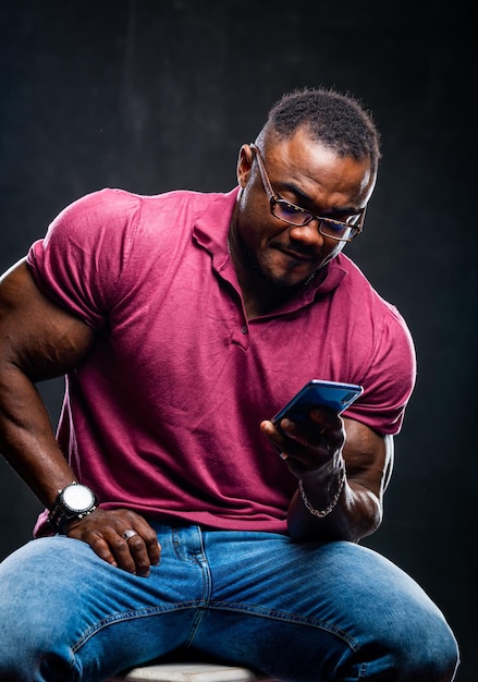 Smiling cheerful young black man poses with phone in hands over black background African american in pink shirt