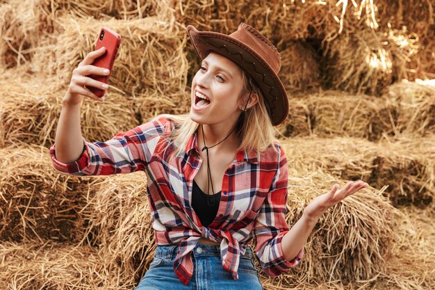 Smiling cheerful young blonde cowgirl sitting on a haystack at the barn, taking a selfie