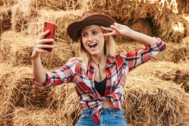 Smiling cheerful young blonde cowgirl sitting on a haystack at the barn, taking a selfie