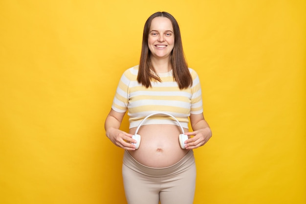Улыбающаяся веселая молодая белая беременная женщина в верхней одежде позирует на ярко-желтой стене с наушниками будущий ребенок слушает классическую музыку для развития