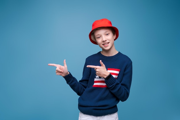 Smiling cheerful young guy in red panama hat and blue sweater with us flag stands pointing with hand