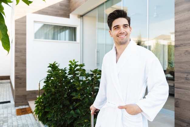 a smiling cheerful young man near hotel outdoors in bathrobe.