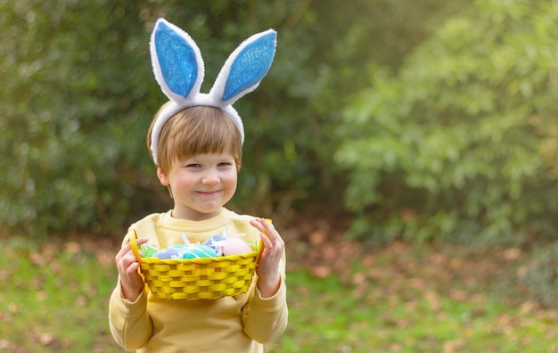 Фото Улыбающийся мальчик с кроличьими ушами и корзиной крашеных яиц в саду пасхальный кролик веселый костюм детства
