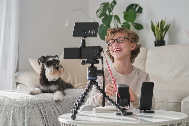 Foto bambino sorridente in abiti casual che registra un video per la trasmissione online mentre è seduto vicino al divano in un salotto moderno con un cane a casa