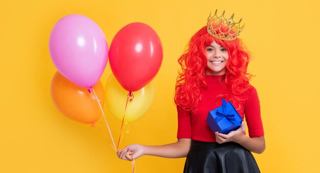 Smiling child in crown with gift box and party balloon on yellow background