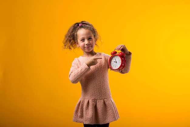 Фото Улыбающаяся девочка держит будильник в руке и указывает на него пальцем концепция образования школа определяет срок обучения