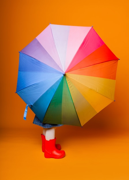 A smiling child girl in a red jacket and boots, holding an\
umbrella, stands on a yellow background. little girl in bright\
clothes holds a rainbow umbrella.