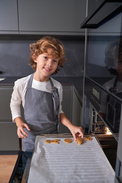 Smiling child in gray apron putting biscuits onto baking paper preparing to cook them in kitchen stove and looking at camera