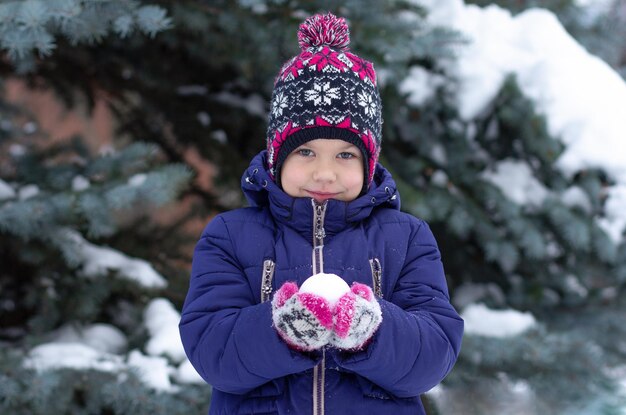 Smiling child holding snowball in hands looking at camera in winter time over snowy spruce backgroun