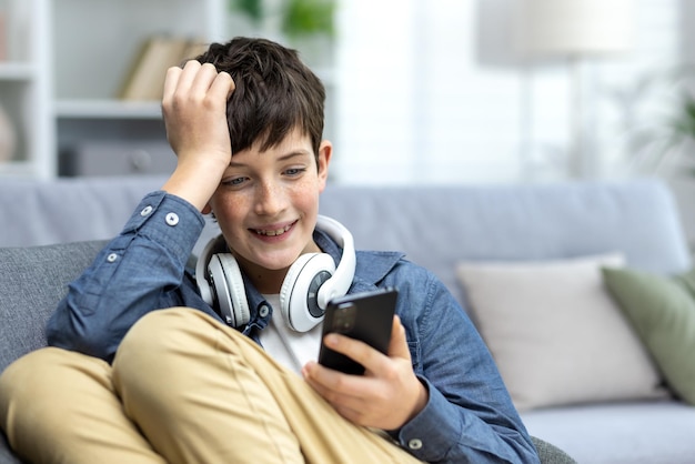 A smiling child of a teenage boy in headphones sitting on the sofa at home resting and using the