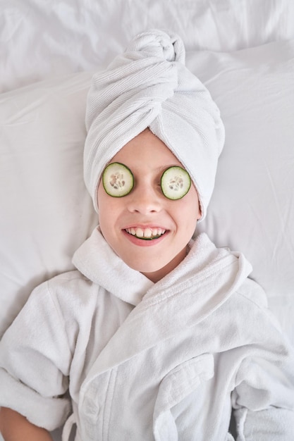 Smiling child with cucumber slices on eyes wearing bathrobe and towel turban relaxing on soft bed during beauty procedures in bedroom at home