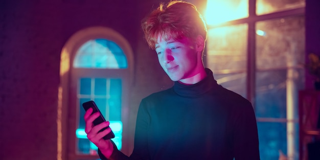 Smiling. Cinematic portrait of stylish redhair man in neon lighted interior. Toned like cinema effects in purple-blue. Caucasian model using smartphone in colorful lights indoors. Flyer.