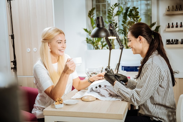 Smiling client. Beautiful blonde-haired client of beauty saloon smiling talking with professional nail artist