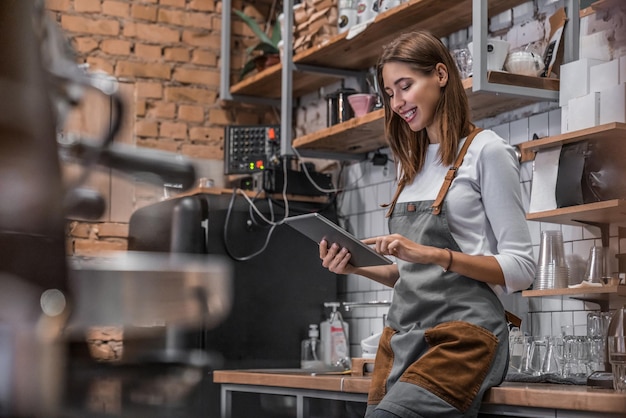 Фото Улыбающаяся владелица кофейни, стоящая на своем рабочем месте возле прилавка и использующая цифровой планшет