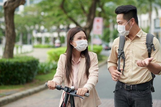 Smiling colleagues in medical masks riding on electric scooters and discussing project details after